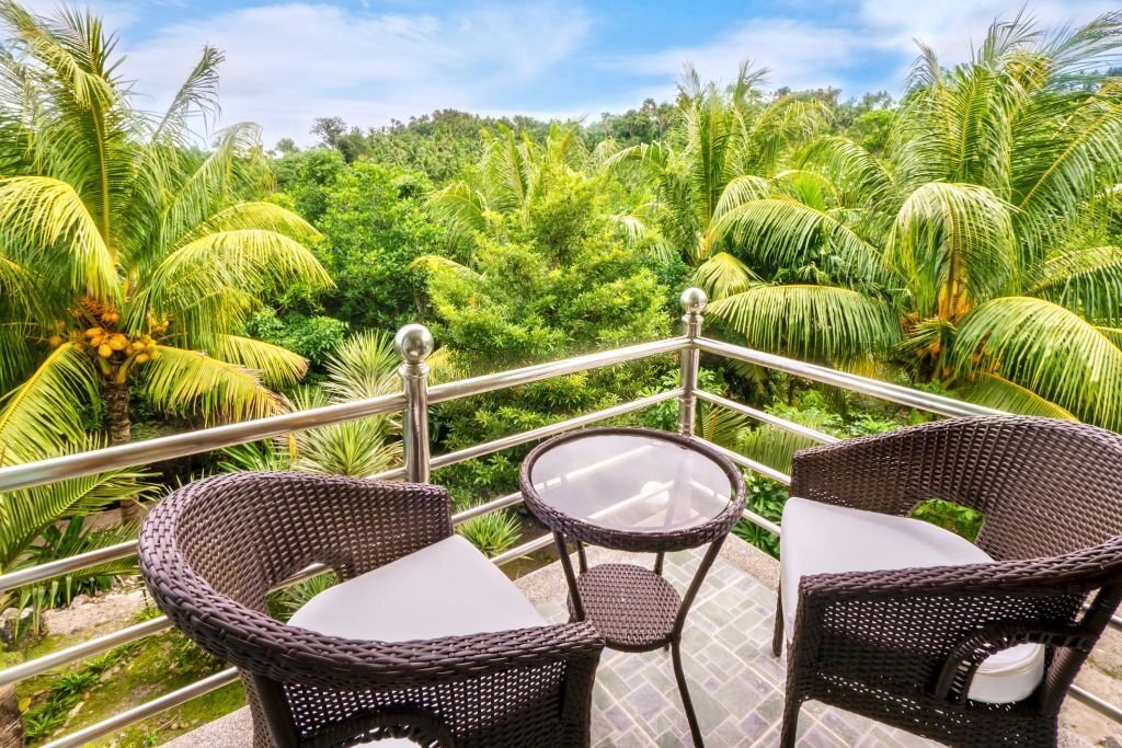 balkon-pribadi A simple balcony set high up, level with a tree canopy in the southeast Asian jungle, with a set of dark brown rattan outdoor furniture, a tiled floor, and shiny chrome railings.