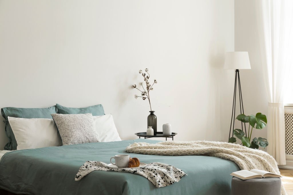 kamar-tidur-luas Bedroom interior with sage green and white sheets and cushions and a blanket. Black metal table with vases beside the bed. A lamp standing in the corner. Real photo.